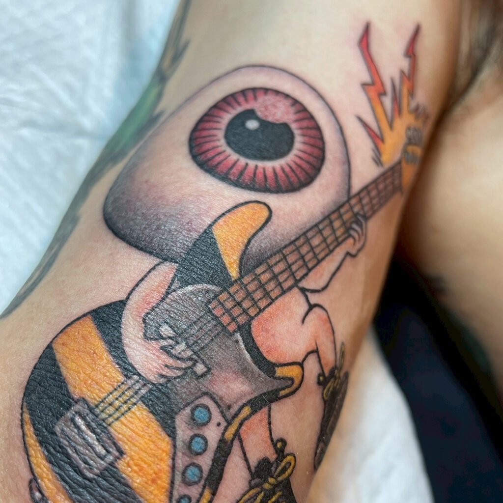 Medama-Oyaji tattoo redesigned to play an electric guitar, featuring a large single eye, tiny body, and bright, bold colors.