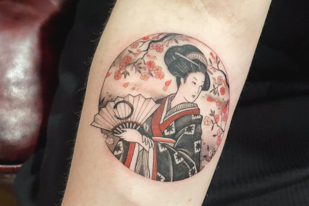Color tattoo of a geisha with fan and cherry blossoms in a circular frame, created at Good Times Ink Osaka.